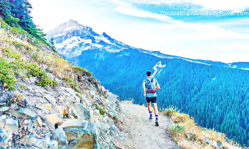 Man in athletic shirt, shorts, and shoes hiking on mountain trail. The beautiful overview is of another mountain with white snowy peaks above and green trees below.